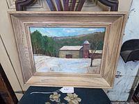Framed unsigned oil painting on board showing barn covered in snow with trees and hills background.