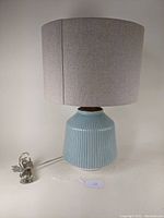 Full view of the table lamp showing the blue ribbed ceramic base, walnut wood neck, light grey fabric drum shade, and attached power cord.