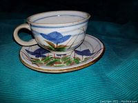 Photo of ceramic teacup and saucer set with blue flowers and green leaves painted on white ceramic base, showing front and side views.