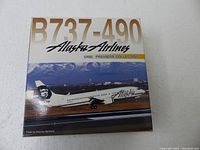 Box front of B737-490 Alaska Airlines 1/400 scale model with photo of airplane on runway, mountains in background. Gold lettering and Alaska Airlines branding.