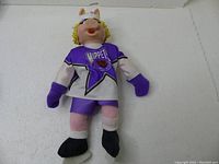 Full view of the vintage Miss Piggy hockey player plush doll in purple and white Muppets jersey lying on a white surface.