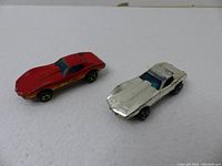 Two Hot Wheels Corvette toy cars, one red with gold stripes and one silver with blue interior, placed on a white background showing overall condition.