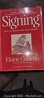 Front cover of a red paperback book titled Signing by Elaine Costello with an image of two hands in a signing pose