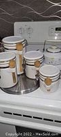 Photo of six white ceramic canisters with attached wooden spoons on a metal stand, plus a white ceramic teapot with lid on a white stove top.