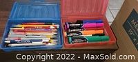 Two open plastic cases displaying markers, pens, and colored pencils inside.