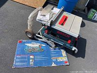 Overall view of table saw on stand with manuals on top and box front on ground