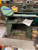 Front view of the green metal cutting bandsaw on stand with manual lying on the stand. Some soiling visible on the machine.