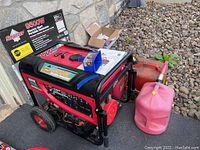 Overall angled view showing generator, manual and funnel on top, original box front behind, two gas cans in foreground
