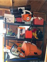 Full shelf showing chainsaw, blower, manuals, gas cans, funnels and fuel