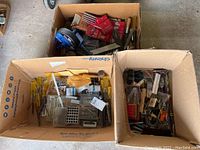 Three boxes containing multiple yellow-handled screwdrivers, files, a blue hand drum auger, and assorted hardware parts
