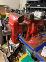 Side angle view of the red King Brand bench vise on a workbench with some workspace clutter around.