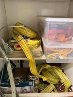 Top shelf of metal cabinet showing yellow ratchet straps and plastic bins containing orange and yellow straps and bungee cords.