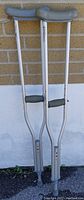 Pair of lightweight silver aluminum adjustable crutches with grey padded underarm rests and hand grips, standing against a brick background.