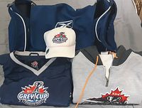 Photo showing the Grey Cup jackets, grey and black hoodie with logos, white Grey Cup hat, and blue Reebok sports bag.