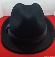 Front angle photo showing the black fedora hat with a black ribbon around the base of the crown on a red background.