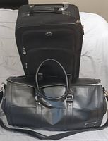 Two carry-on bags: black wheeled suitcase standing upright and black duffle bag in front on bed.
