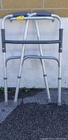 Front view of silver aluminum walker with blue padded handles and rubber leg tips against a brick wall.