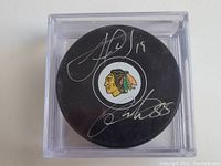 Black hockey puck with Chicago Blackhawks logo signed by Jonathan Toews (19) and Patrick Kane (88) in silver ink, displayed in clear protective case.