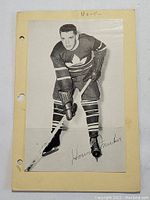 Front view of black and white photo print of Howie Meeker in hockey gear on beige paper with three-hole punches.