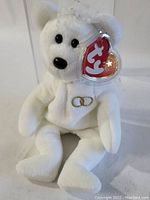 White TY Beanie Baby "Valentino" bear with gold embroidered rings on chest, heart-shaped TY tag with star, photographed outside of acrylic case.