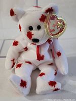 Front view of white plush bear with red maple leaf patterns and heart-shaped Ty tag on ear, placed inside clear display box.