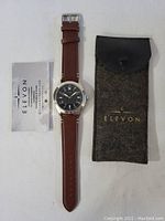 Full view of the Elevon watch lying flat with brown leather strap, alongside its original felt pouch and warranty card on white background.