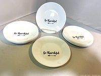 Set of twelve Corelle dessert plates arranged in three stacks with one plate displayed upright on a stand, showing the central 'be thankful' text and arrow design.