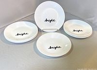 One dozen white dessert plates displayed with the black script 'bright' in the center