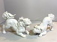 Pair of white porcelain foo dogs shown side by side from side angle, showcasing detailed design and size relation.