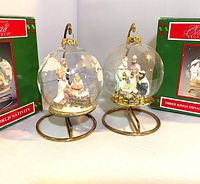 Pair of two House of Lloyd Christmas ornaments featuring nativity scenes inside frosted glass globes on gold-tone metal stands with original boxes visible behind.