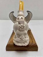 Front view of pipe showing detailed carved head of a man with beard, turban, and flower on top. Pipe is on wooden display stand with carved upright supports seen in background.