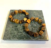 Two tiger eye tumble bead bracelets displayed on a greenish marble square tile showing their size and smooth polished finish.