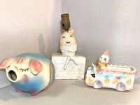 Three vintage ceramic baby decor items: piggy bank, Humpty Dumpty lamp, and ice cream truck container on white background.