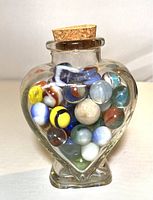 Heart-shaped glass bottle filled with assorted vintage clay and glass marbles, sealed with a cork stopper, showing multiple angles of the marbles and bottle shape.