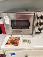 Front view of the stainless steel pressure oven with product guide and recipe book placed in front of it on a white countertop.