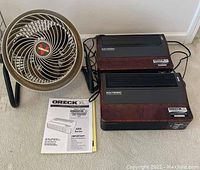 Two Oreck XL Air8 Series professional air purifiers, one Oreck manual, and one Vornado floor fan shown on carpeted floor near white wall.