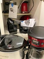 Kitchen shelf with Hamilton Beach slow cooker (silver with glass lid), Mr. Coffee coffee maker (red and black), Bella waffle maker (black), Oster toaster, Oster hot plate, Presto electric multi cooker, red Nostalgia popcorn popper and manuals.