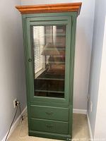 Front view of tall green media cabinet with glass door showing shelves inside and two bottom drawers