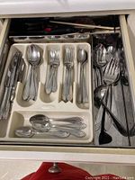 Drawer with flatware set including knives, forks, spoons in a plastic organizer, with additional serving spoons and forks on metal tray.