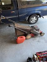 Full side view of Snapper lawnmower with attached grass bag and red gas can on concrete garage floor next to a vehicle.