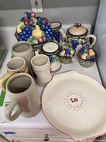 Full view of the Mikasa Garden Harvest ceramic set including cookie jar, candleholders, teapot, creamer, and sugar bowl alongside four ceramic beer steins and Salem Calico serving plate.