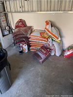 Photo showing several large bags of planting materials stacked against a garage wall including mulch and various soil bags.
