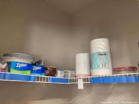 Shelf with paper plates, Ziploc sandwich and freezer bags, plastic food storage containers, paper towels, coffee filters, and trash bags arranged in original packaging.