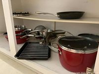 Photo of the set of pots and pans on a shelf showing red exterior, metal handles, glass lids, broiling rack, and rack insert.