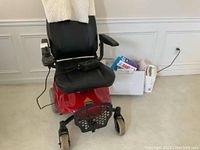 Front view of red Shoprider power chair with black seat, footrest, front wheels and control module; box of bandages visible at right