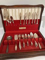 Opened wooden flatware box showing red velvet interior holding assorted silver plate flatware including forks, spoons, and serving spoons with ornate handle designs