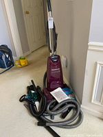 Photo showing full upright vacuum cleaner with flexible hose, several attachments, vacuum bag and instruction manual placed at the base and around the vacuum.