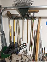 Photo showing various hanging yard tools including shovels, rakes, and pruners attached to wall rack; garden hose coiled in spreader cart on floor.