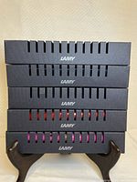 Photo shows five stacked LAMY Safari fountain pen boxes with ventilation slits revealing pen colors including red and pink, displayed on a small stand against a beige textured background.