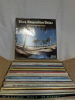 Photo showing a collection of over 50 vinyl records in a cardboard box including 'Blue Hawaiian Skies' by Big Ben Hawaiian Band at top, all are previously owned and appear in good condition.